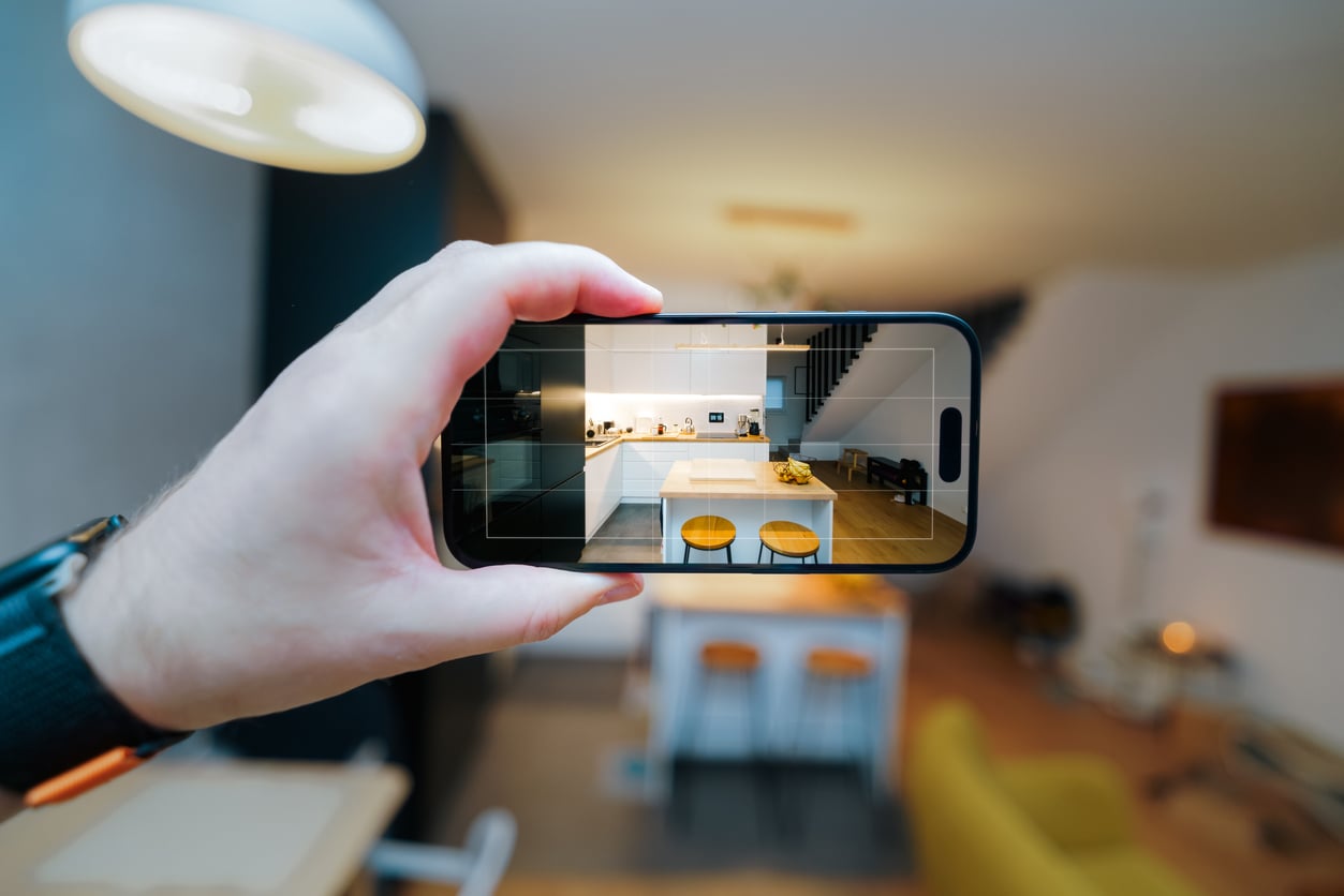 A person holds a smartphone, capturing a photo of a modern kitchen with white cabinets, wooden countertops, and two stools—showcasing the inviting space while staying at an Airbnb. The phone screen displays the kitchen's interior in clear focus.