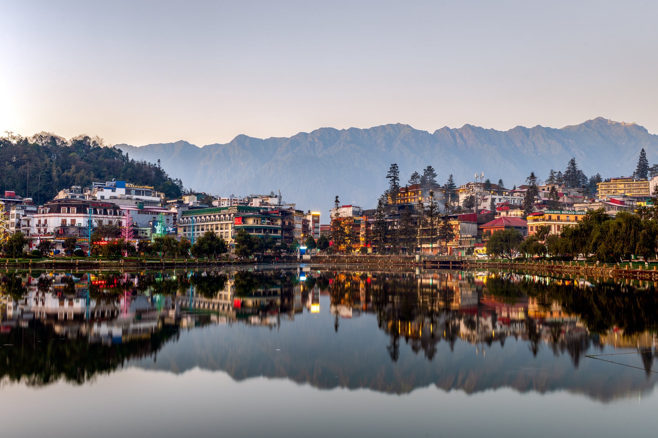 Colorful buildings and trees line the edge of a calm lake, reflecting perfectly in the water. Majestic mountains rise in the background under a clear sky, creating a peaceful and picturesque scene.