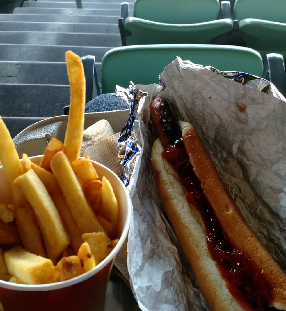 A cup of golden French fries sits beside a fast-food hot dog topped with ketchup, wrapped in foil, with stadium seats and steps visible in the background.