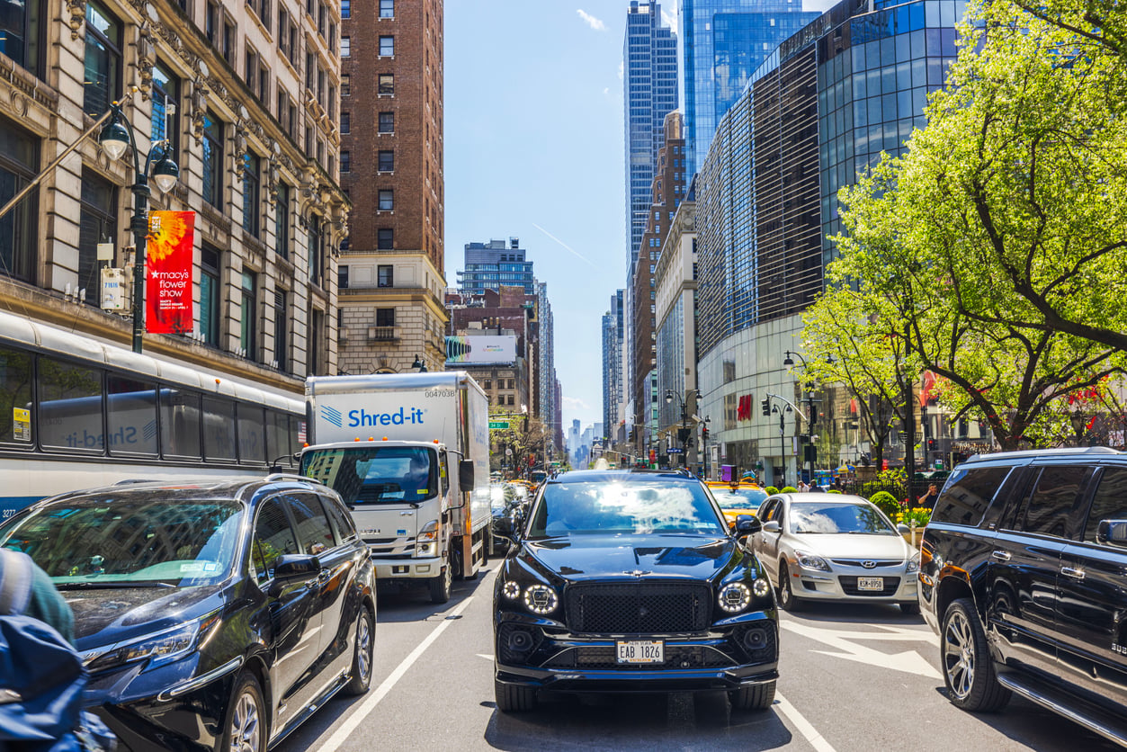 Busy city street with delayed purchases evident as vehicles, including a black SUV, are stopped in traffic. Multiple cars and trucks fill the road, tall buildings rise on both sides, and trees line the sunny sidewalks under a clear sky.