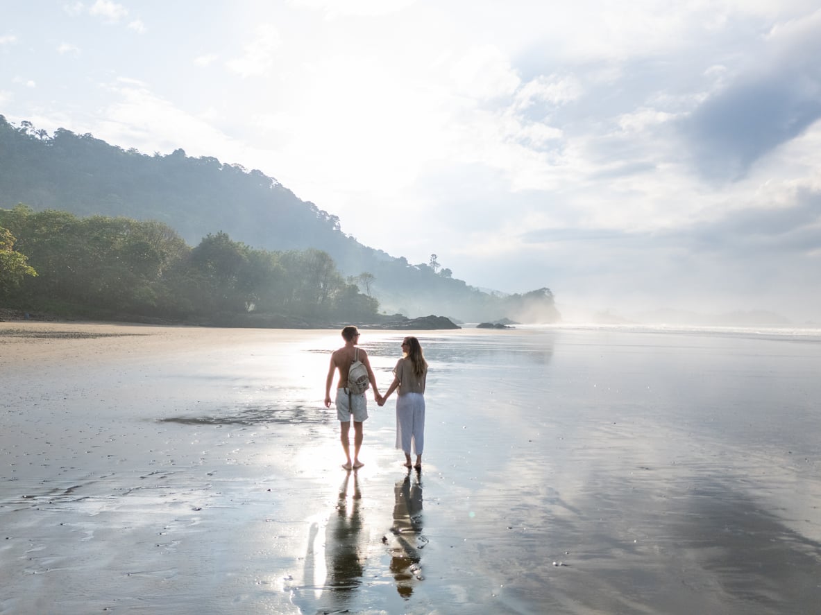 A couple holding hands walks along a calm, reflective beach—one of the serene Florida alternatives—with misty hills and trees in the background under a cloudy sky.