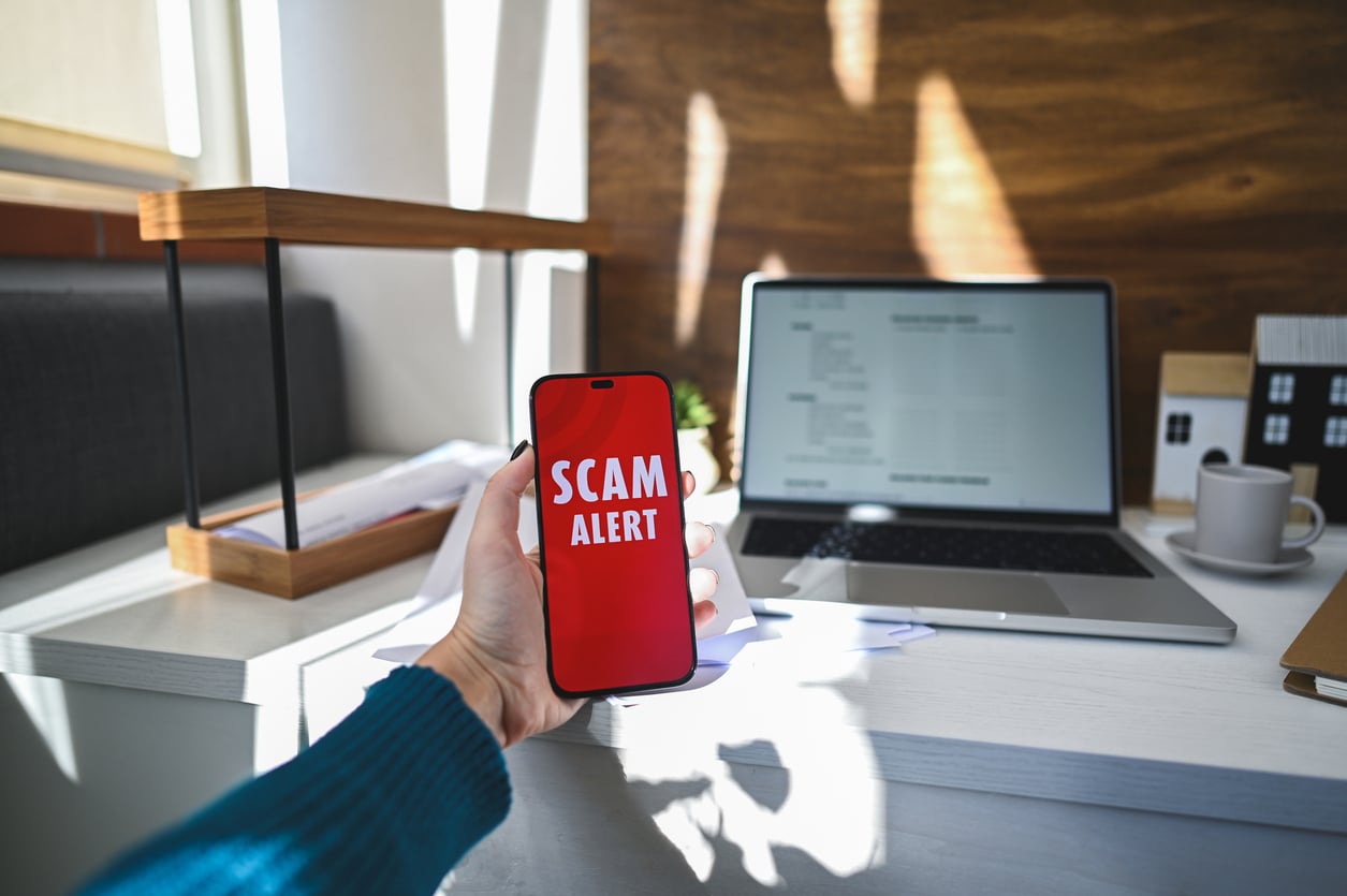 A person staying at an Airbnb holds a smartphone displaying a red screen with the words “SCAM ALERT” in a home office, with a laptop, desk lamp, and documents in the background.