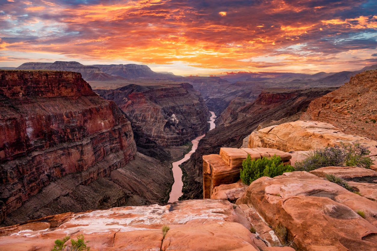 A dramatic sunset illuminates the Grand Canyon, one of the world’s top tourist attractions, highlighting its steep red rock cliffs and the winding Colorado River below as vibrant orange, pink, and purple clouds fill the sky.