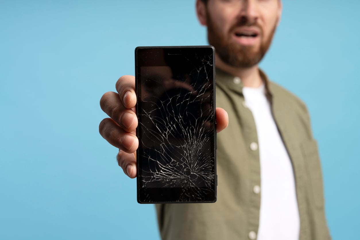A person with DIY skills holds out a smartphone with a badly cracked screen, displaying frustration. The background is plain blue, and the person wears a green shirt over a white t-shirt.