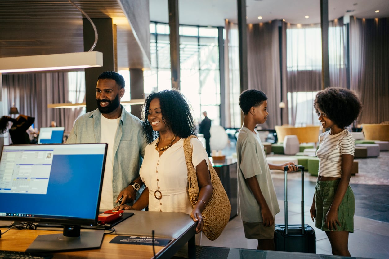 A family of four stands at a hotel reception desk checking in, their smiles hinting at excitement for upcoming adventures. The parents chat with the front desk while their children, armed with luggage and theme park hacks, wait in the modern, well-lit lobby.