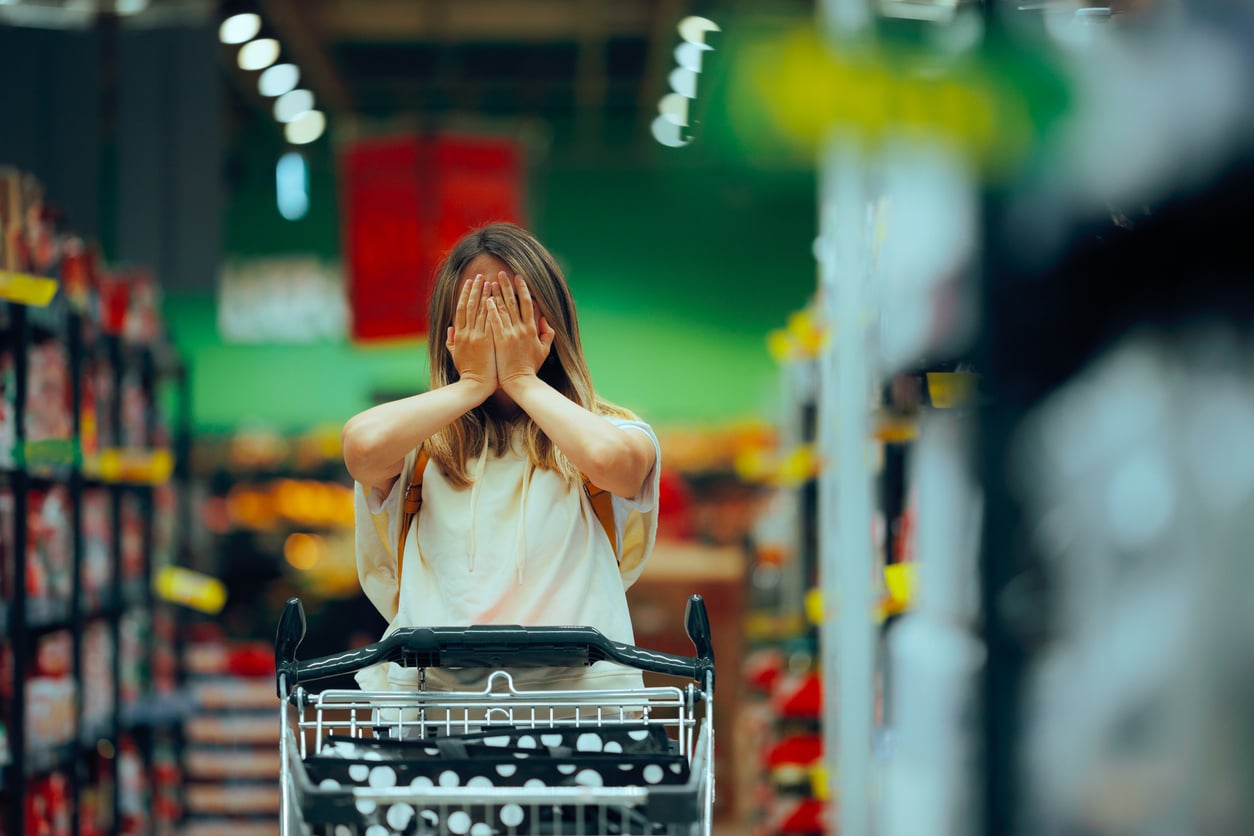 A person stands in a supermarket aisle, covering their face with both hands while holding onto a shopping cart, surrounded by shelves stocked with various products.