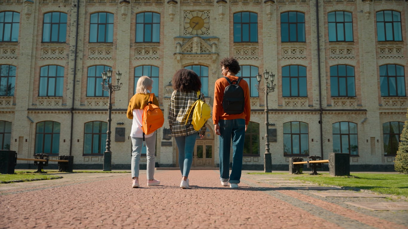 Three students with backpacks walk towards a large, historic university building with tall windows and a clock above the entrance, on a sunny day.