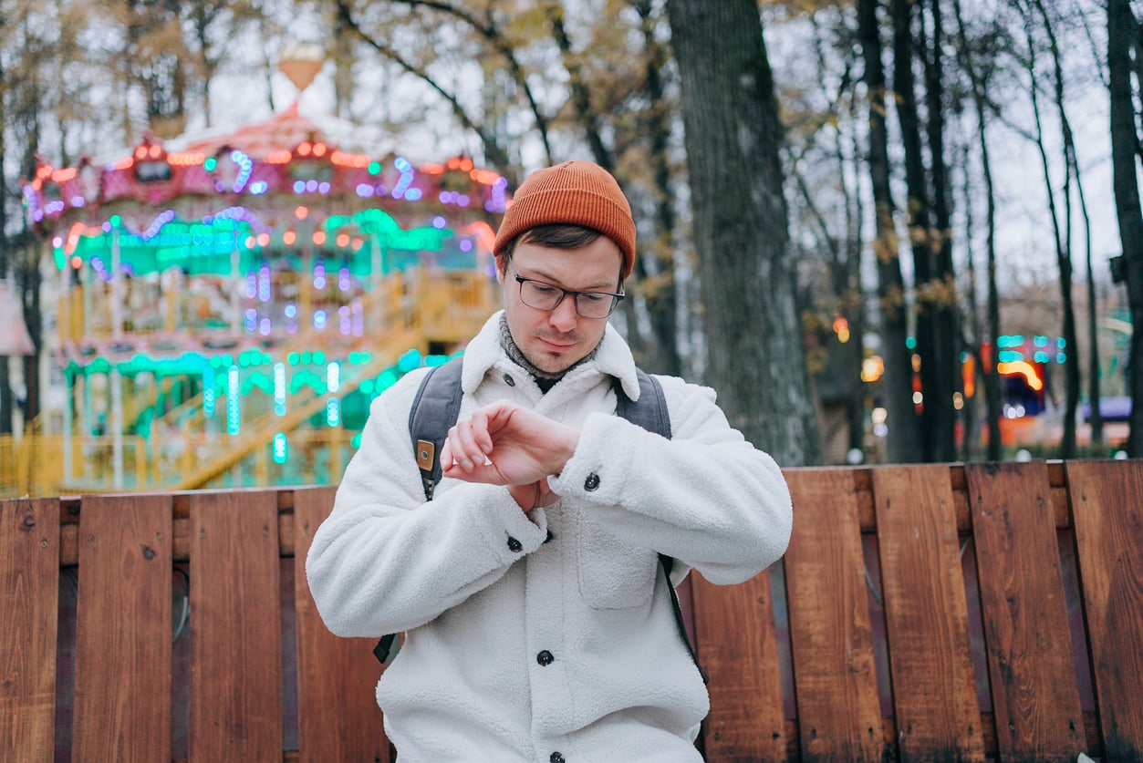 A man in a white jacket and orange beanie looks at his watch while sitting on a wooden bench, perhaps planning his next theme park hacks. Behind him, colorful lights from a carousel glow among trees in the park.