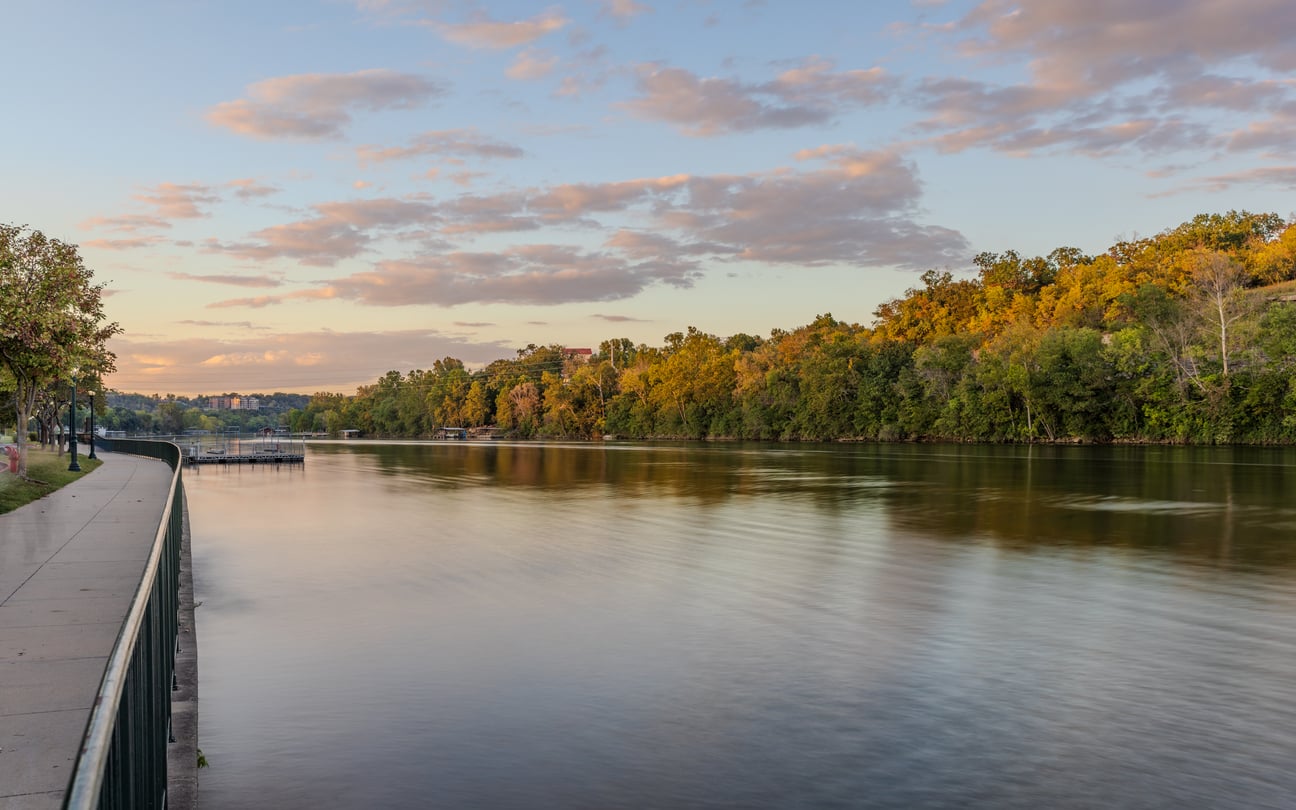 A calm river flows alongside a tree-lined walkway under a partly cloudy sky at sunset, with golden light illuminating the foliage and reflecting on the water—showcasing one of many serene Florida alternatives for peaceful evening strolls.