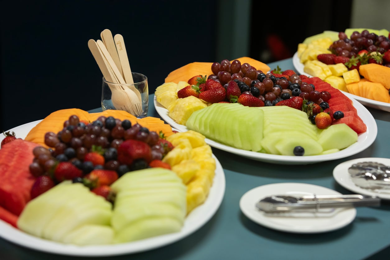 Three large platters of assorted fresh fruit, including cantaloupe, watermelon, honeydew, strawberries, grapes, blueberries, and pineapple. Wooden utensils are in a glass, with serving tongs and plates nearby on the table.