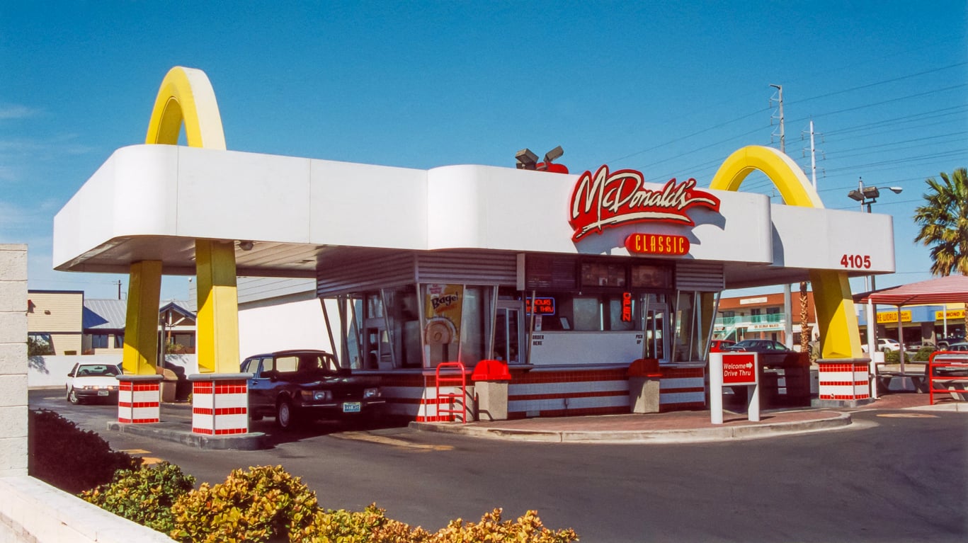 A retro-style McDonald's restaurant with yellow arches, a red "McDonald's Classic" sign, and a drive-thru captures the charm of Vintage Fast-Food Photos. A vintage car is parked outside, framed by palm trees and shops under a clear blue sky.