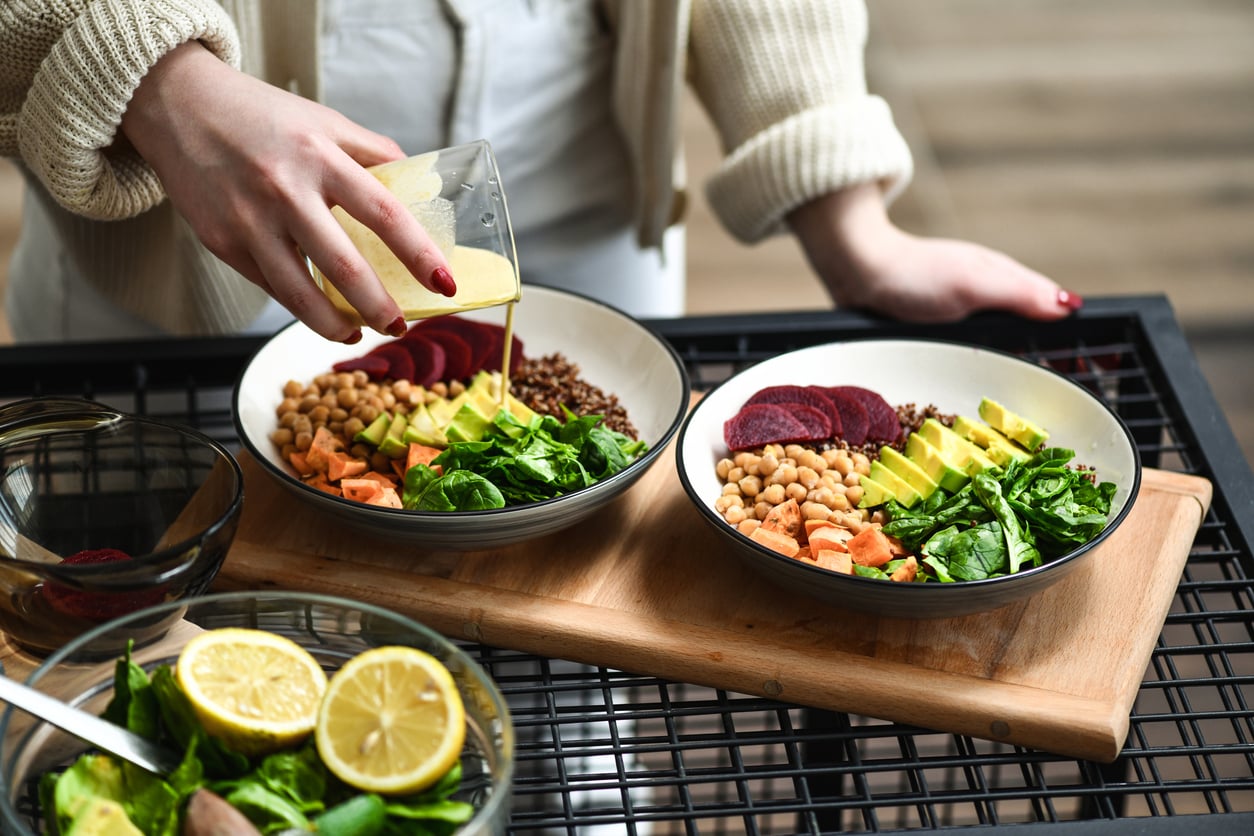 A person pours dressing onto a bowl filled with avocado, chickpeas, spinach, sweet potatoes, beets, and grains—delicious ground beef alternatives. Another similar bowl and halved lemons are on the table.
