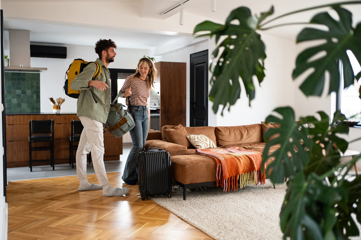 A man and woman with backpacks and luggage smile as they enter a modern, cozy living room with a brown sofa, green plant, and wooden floor. The kitchen is visible in the background.