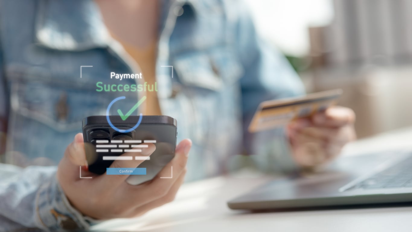 A person holds a smartphone and a credit card, with a laptop nearby. On the image, a graphic overlay displays “Payment Successful” with a checkmark and digital payment interface elements.