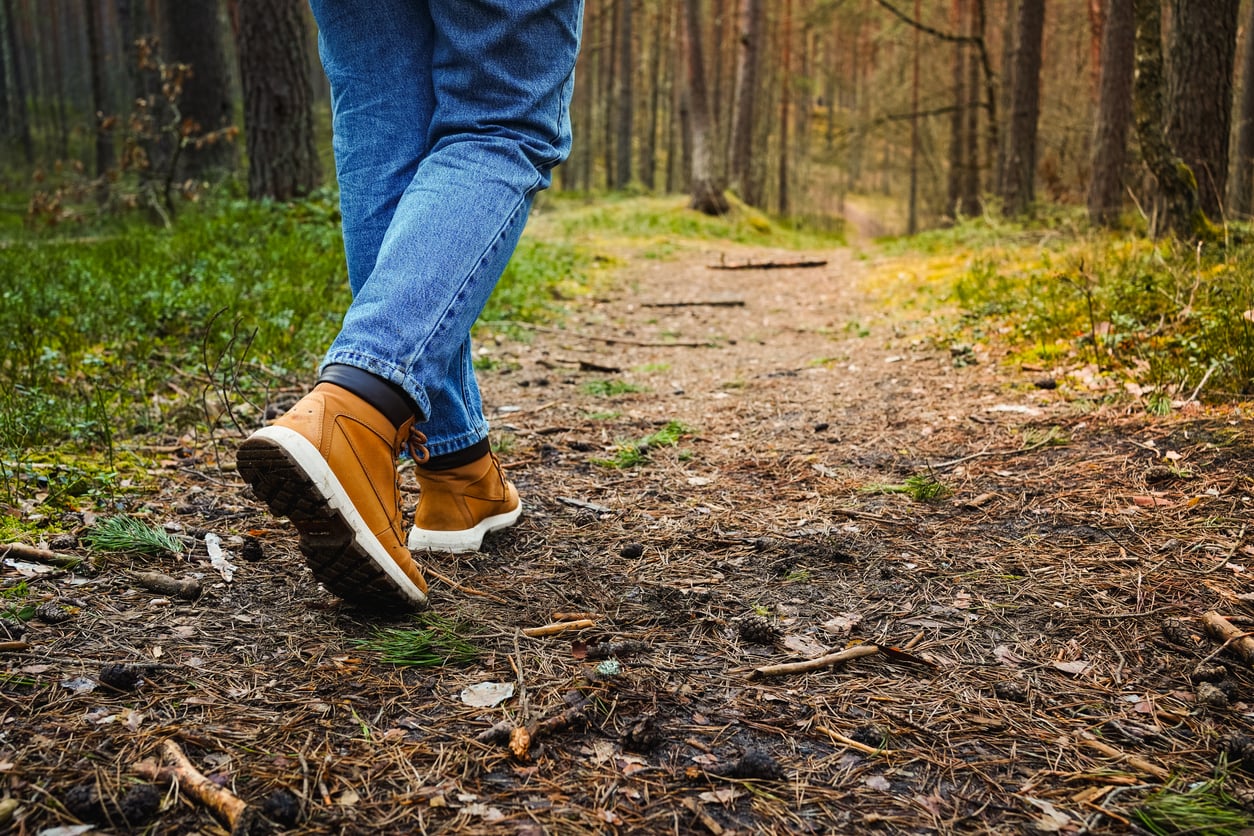 Person wearing blue jeans and tan hiking boots walking along a dirt path through a forest, surrounded by green grass and tall trees—a peaceful outdoor walk that supports mental health and relaxation.