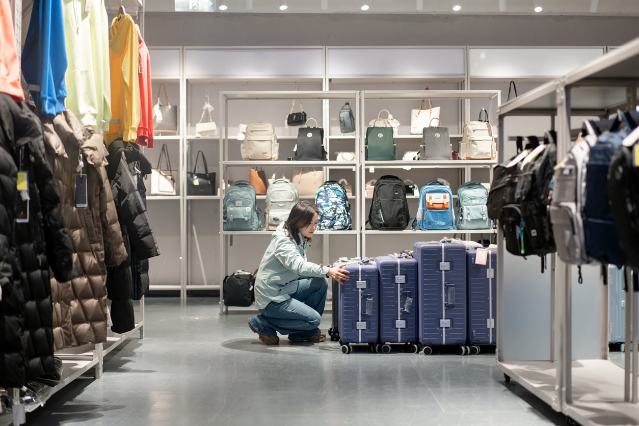 A person crouches in a store aisle, examining a stack of blue suitcases. Shelves around them display backpacks, jackets, and handbags. The store is well-lit and neatly organized.