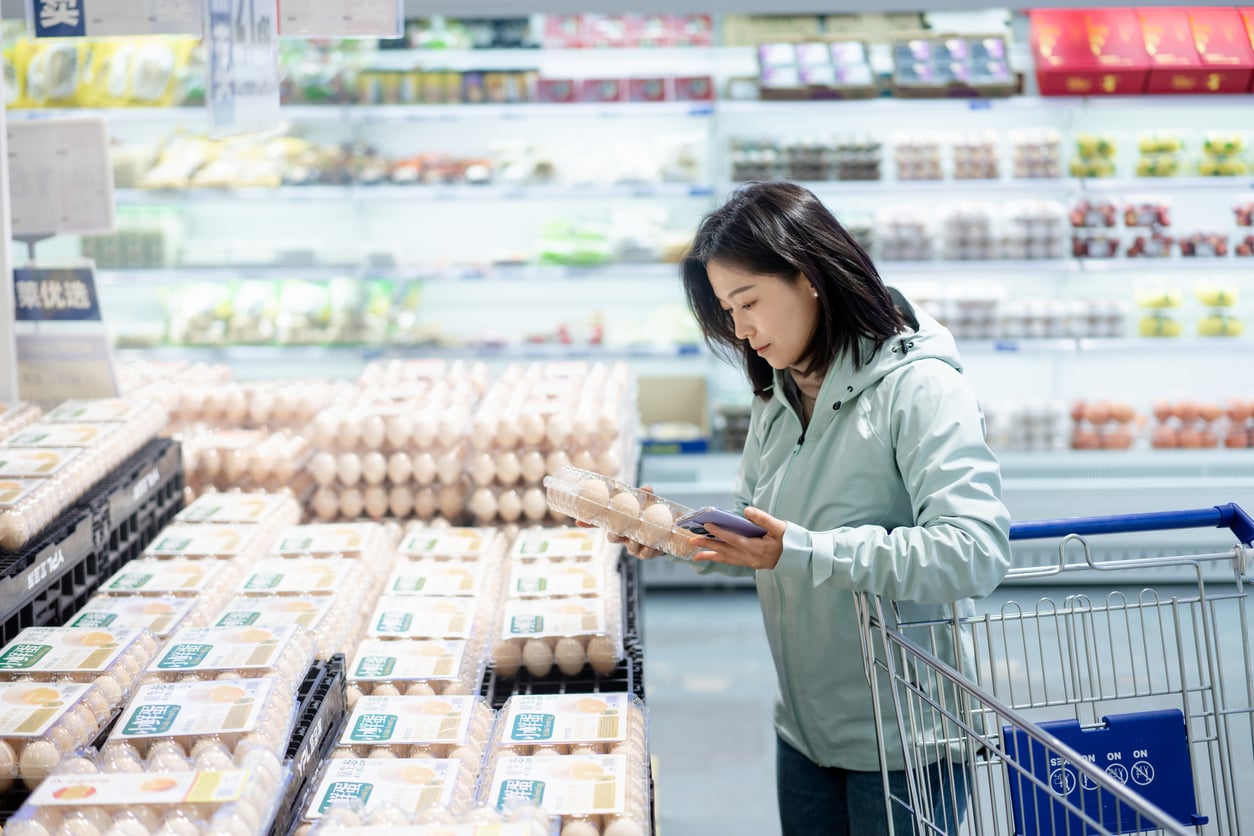 A woman in a light jacket stands by a shopping cart in a supermarket, examining a carton of eggs among stacks of overpriced products and other food items in the refrigerated section.