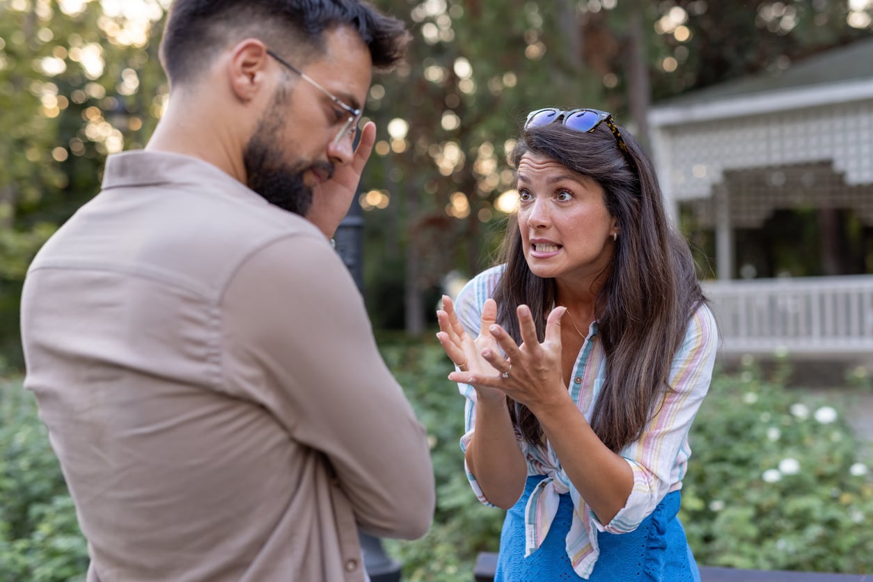 A woman with long hair gestures emotionally while talking to a man with a beard who stands with his arms crossed, looking away. They appear to be having an intense conversation outdoors in a garden setting.