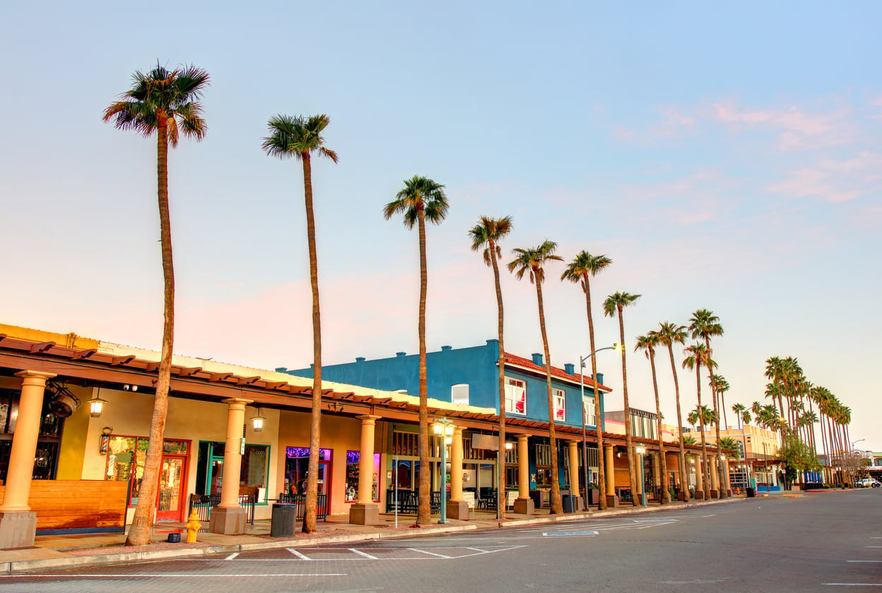 A row of tall palm trees lines the street in front of colorful buildings with shops and restaurants at sunset, under a clear sky. The scene is quiet with no people or cars present.