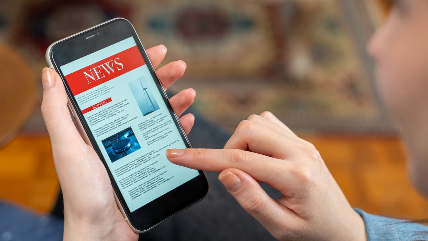 A person holding a smartphone and reading an online news article about mental health, with images and text visible on the screen against a blurred indoor background.