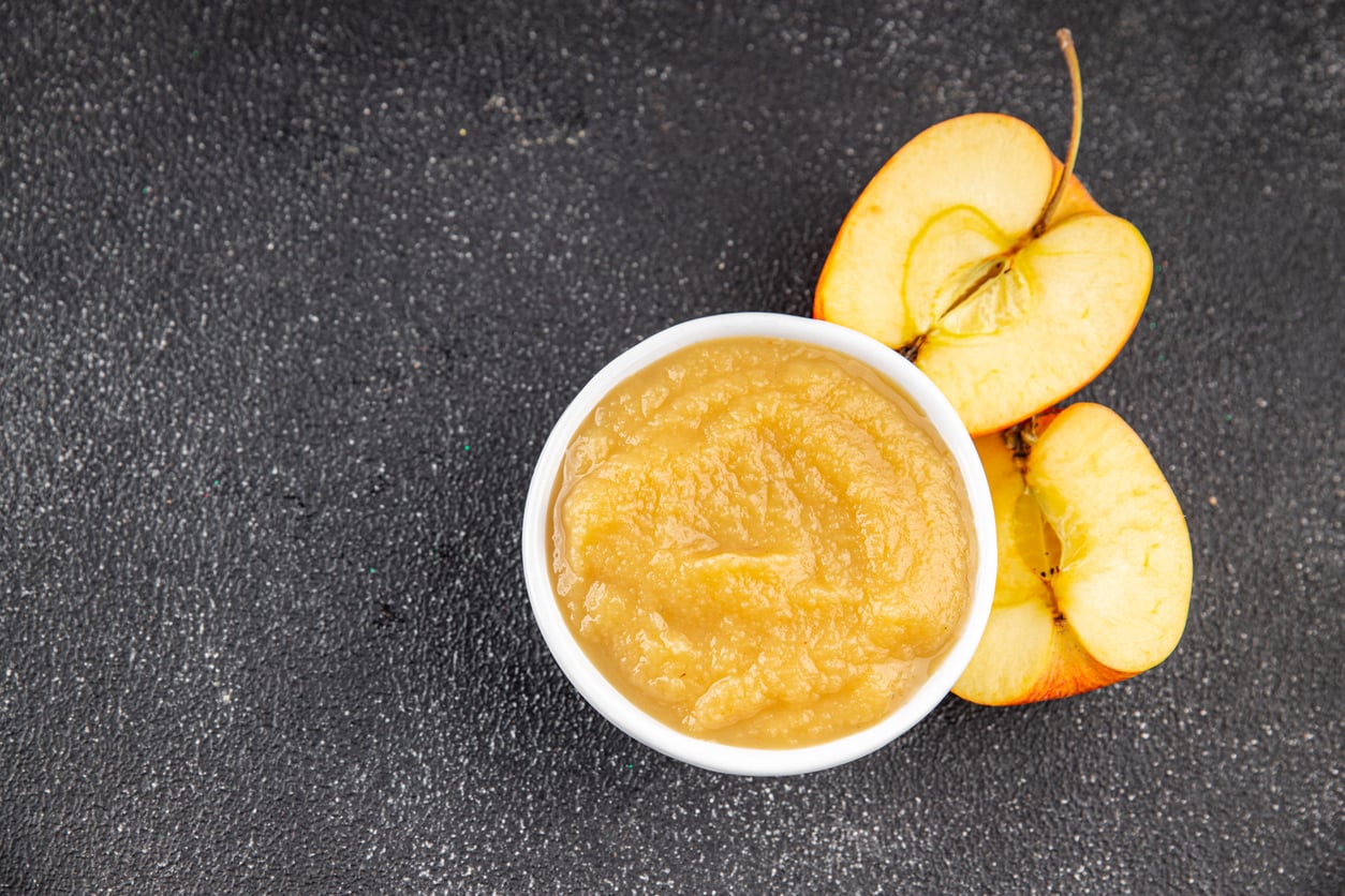 A small white bowl filled with applesauce, a popular egg substitute, sits beside two apple slices on a dark textured surface.