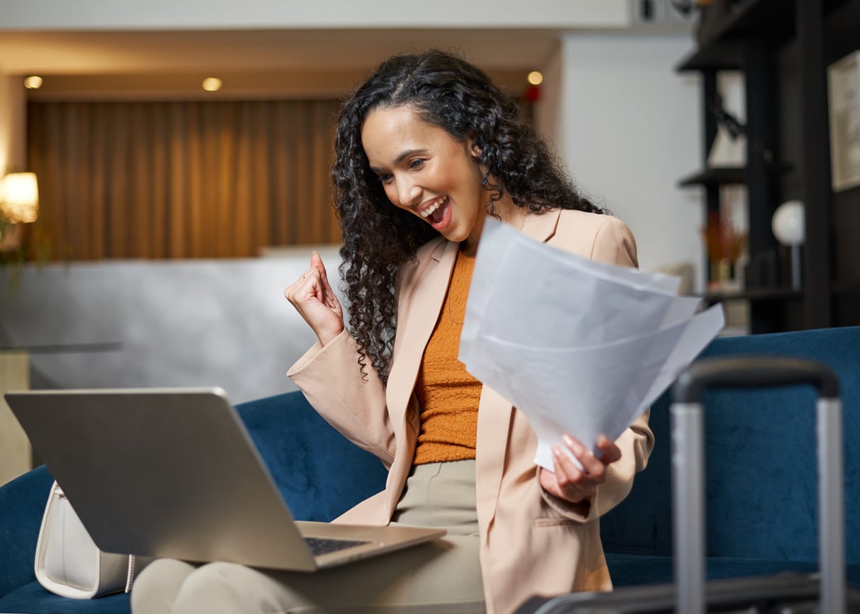 A woman sitting on a blue sofa looks excited while looking at her laptop, holding papers in one hand and making a fist pump with the other. A suitcase is visible in the foreground, suggesting a travel setting.