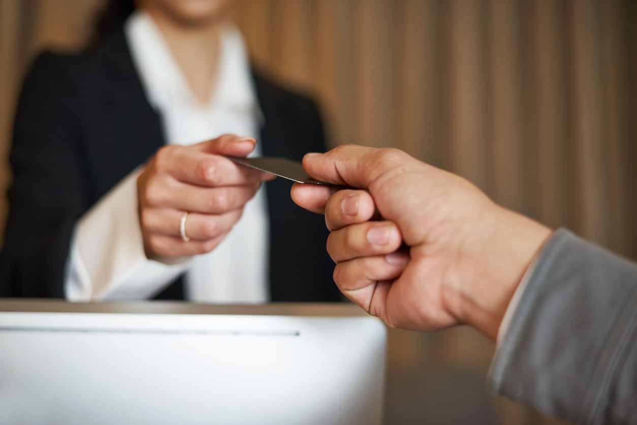 A person in a suit hands a key card to another over a reception desk, suggesting a check-in or service interaction. The faces are out of focus, emphasizing the exchange and highlighting Hotel Room Safety during the process.