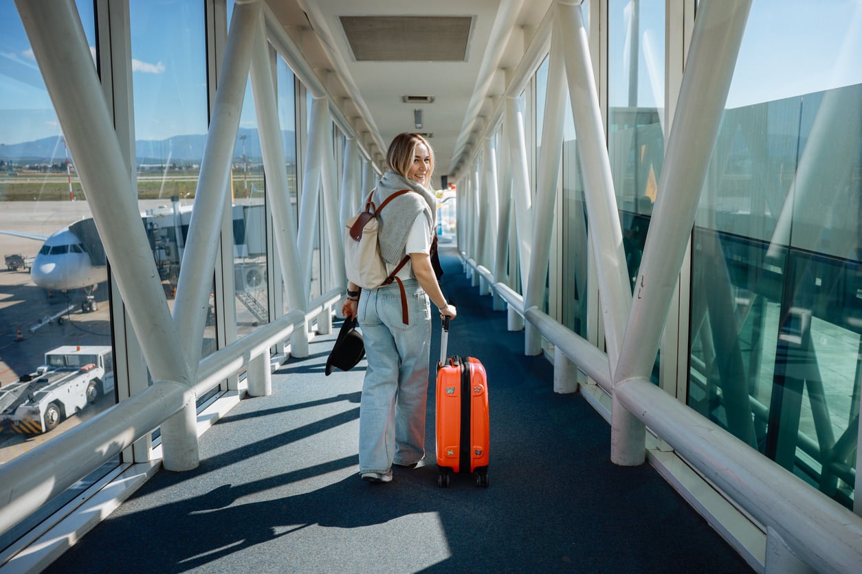 A person with a backpack and orange suitcase walks down an airport jet bridge toward an airplane, looking back and smiling. Sunlight streams through large windows, and the runway is visible outside.