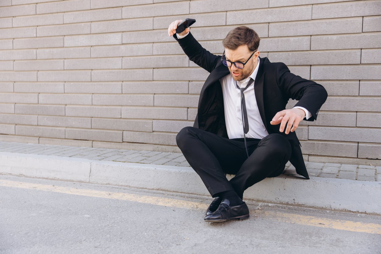 A man in a suit and glasses sits on the curb, looking frustrated as he drops his phone—perhaps after missing out on concert ticket hacks—with one arm raised and an upset expression, against a brick wall background.