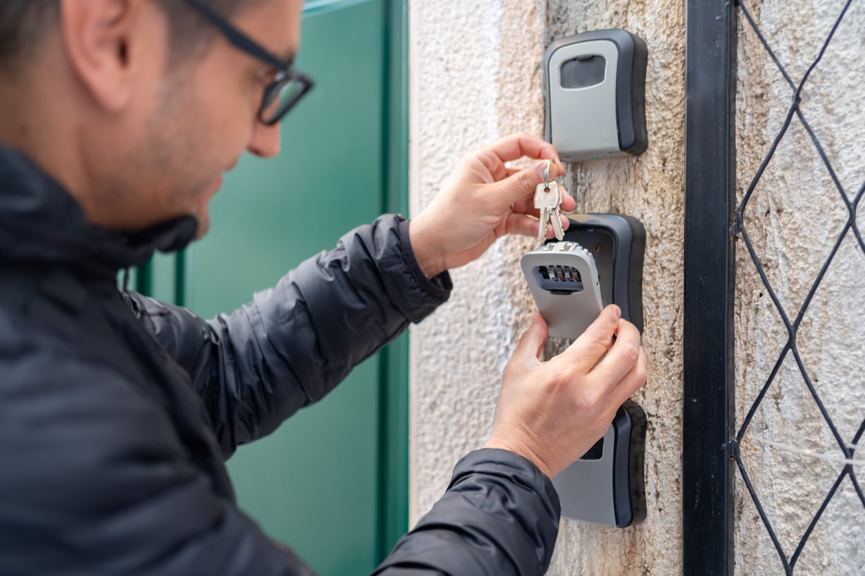 A person wearing glasses and a black jacket is placing keys into a wall-mounted lockbox beside a green door and a wire fence.