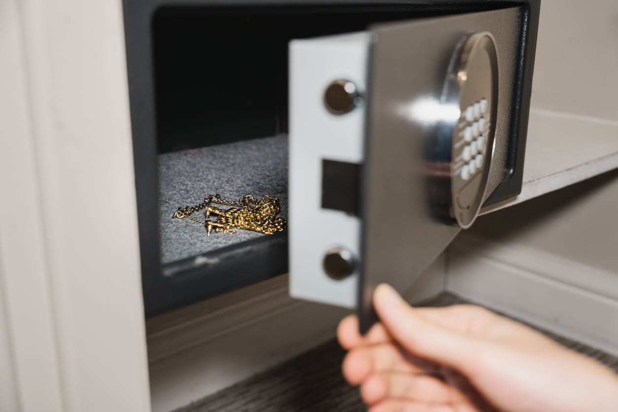 A hand opens a metal safe with an electronic keypad inside a hotel room, revealing gold jewelry on the safe's floor—showcasing reliable hotel room safety for your valuables.