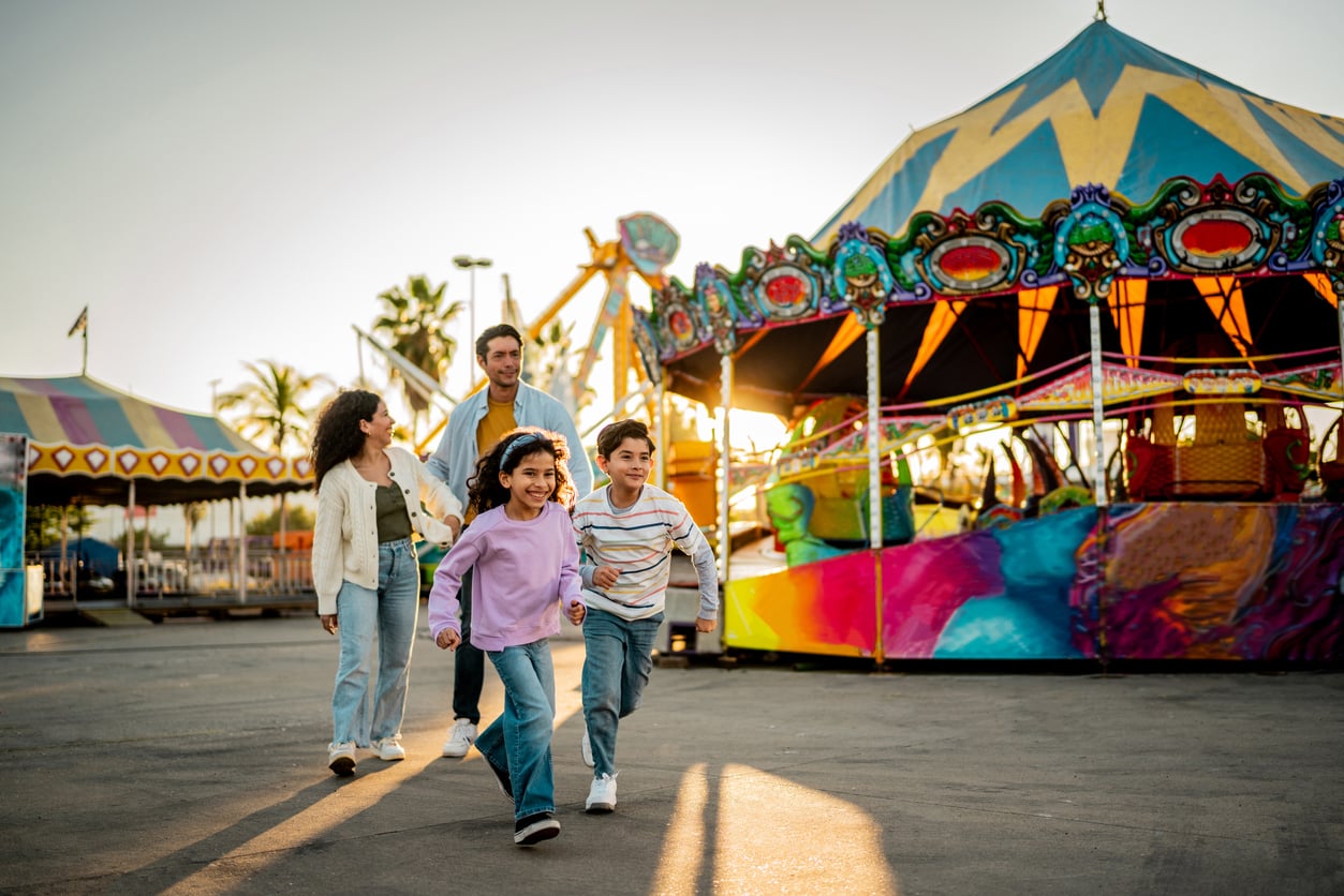 A family of four enjoys a day at an amusement park, with two children running ahead and two adults following behind, while discovering theme park hacks. Colorful carnival rides and a merry-go-round brighten the background.