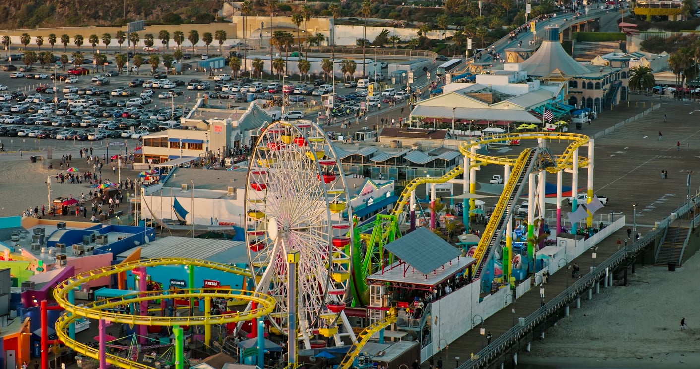 Aerial view of a busy amusement park on a pier, featuring a large Ferris wheel, colorful roller coaster, various rides, and people walking—perfect for discovering theme park hacks to make the most of your visit. Nearby parking lot filled with cars.