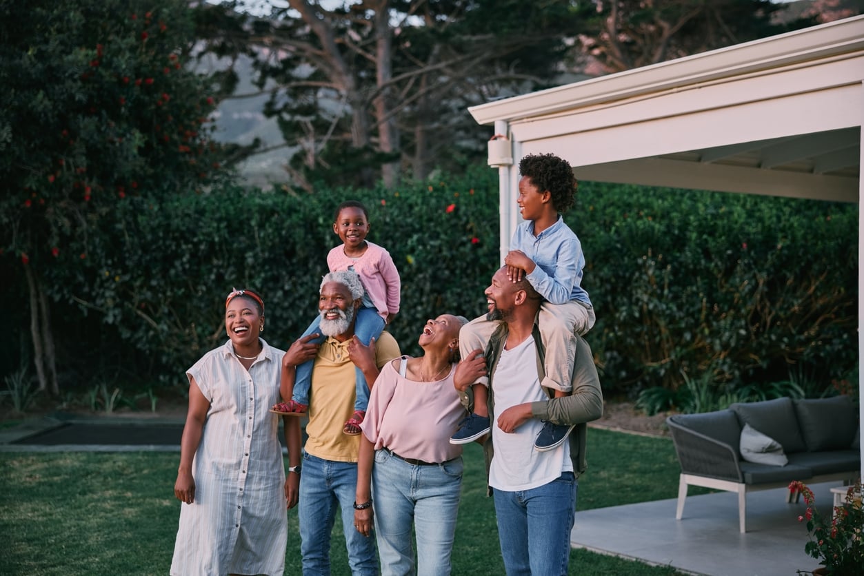 A multigenerational family of six, including two children, stands outdoors in a backyard. They are smiling and laughing, with one child on an adult’s shoulders, enjoying time together near a porch and greenery.
