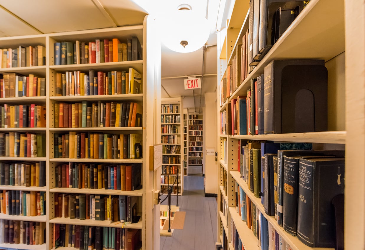 Bookshelves filled with colorful books line a narrow hallway in a library or bookstore, offering titles about Small Business States. An exit sign glows above an open doorway leading to more shelves, while warm lighting brightens the inviting scene.