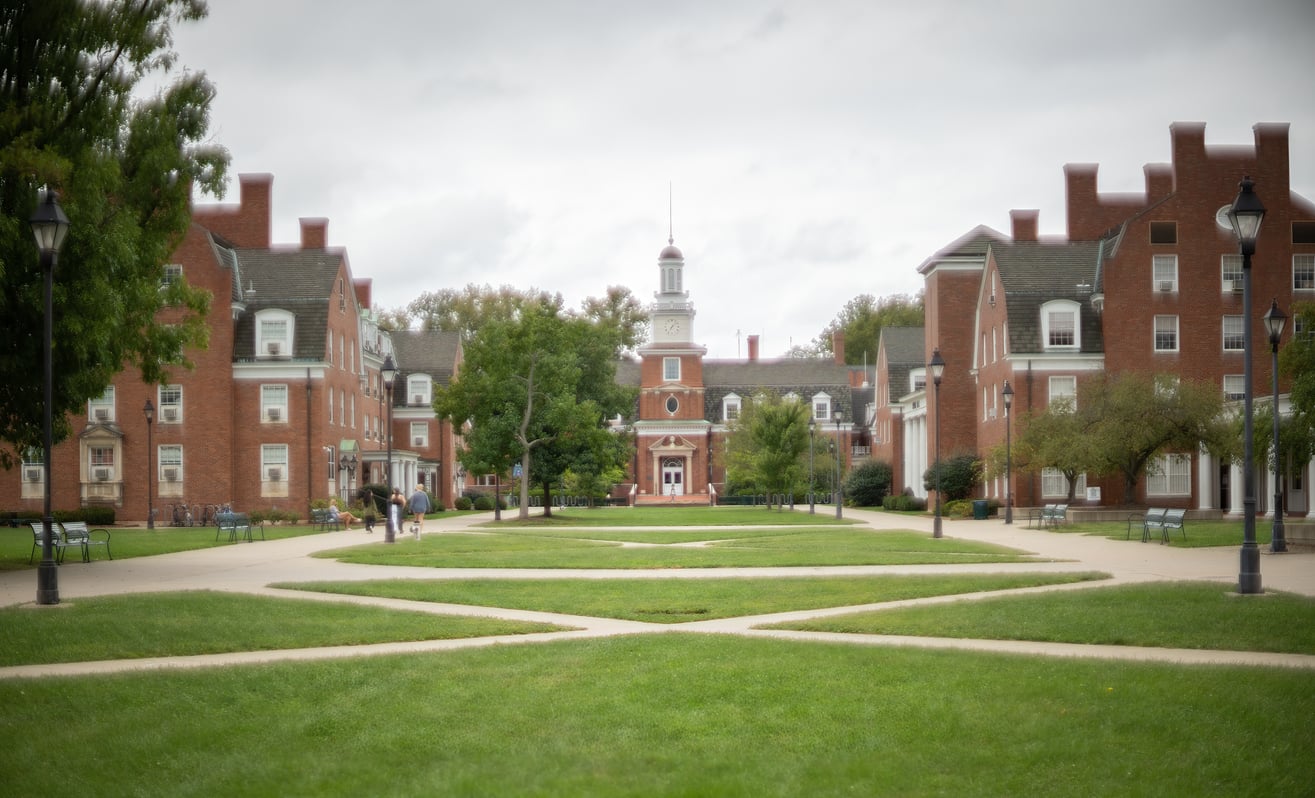 A grassy quad with crisscrossing sidewalks is surrounded by red-brick academic buildings in one of the most affordable college towns, with a white clock tower at the center under a cloudy sky. A few people walk along the paths.