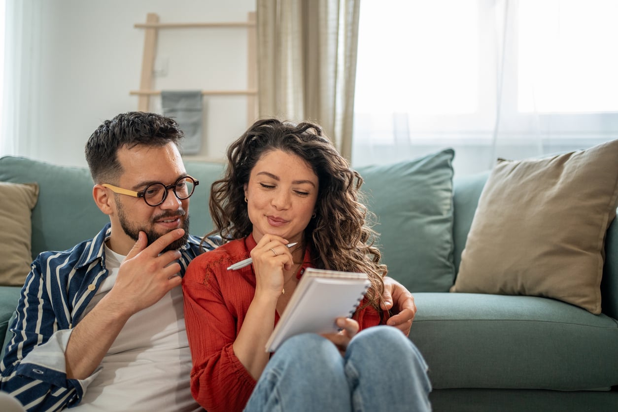 A couple sits on the floor in front of a sofa, looking thoughtful. The woman holds a notepad and pen, appearing to write, while the man sits close beside her, resting his hand on his chin.