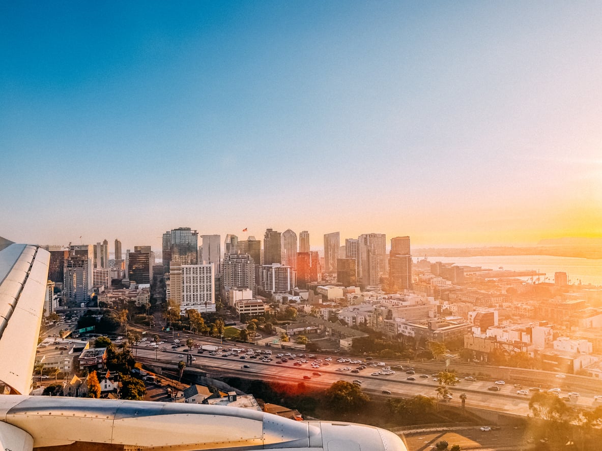 View from an airplane window approaching a city skyline at sunset, with tall modern buildings, highways, and traffic below—sunlight illuminating the scene near some of the city's most convenient airports.
