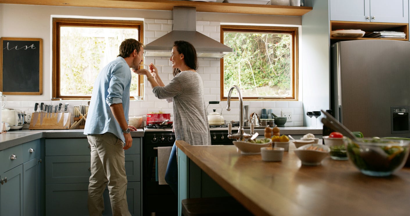 A woman playfully feeds a man with a spoon in a bright, modern kitchen. They are standing near a stove, smiling, with various kitchen items and bowls on the counters around them.