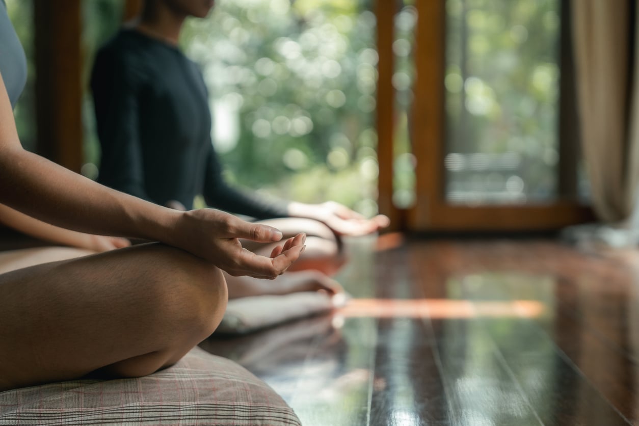 Two people sit cross-legged indoors on cushions in a meditative pose, hands resting on knees with palms up, practicing mindfulness for mental health in front of large windows with greenery visible outside.