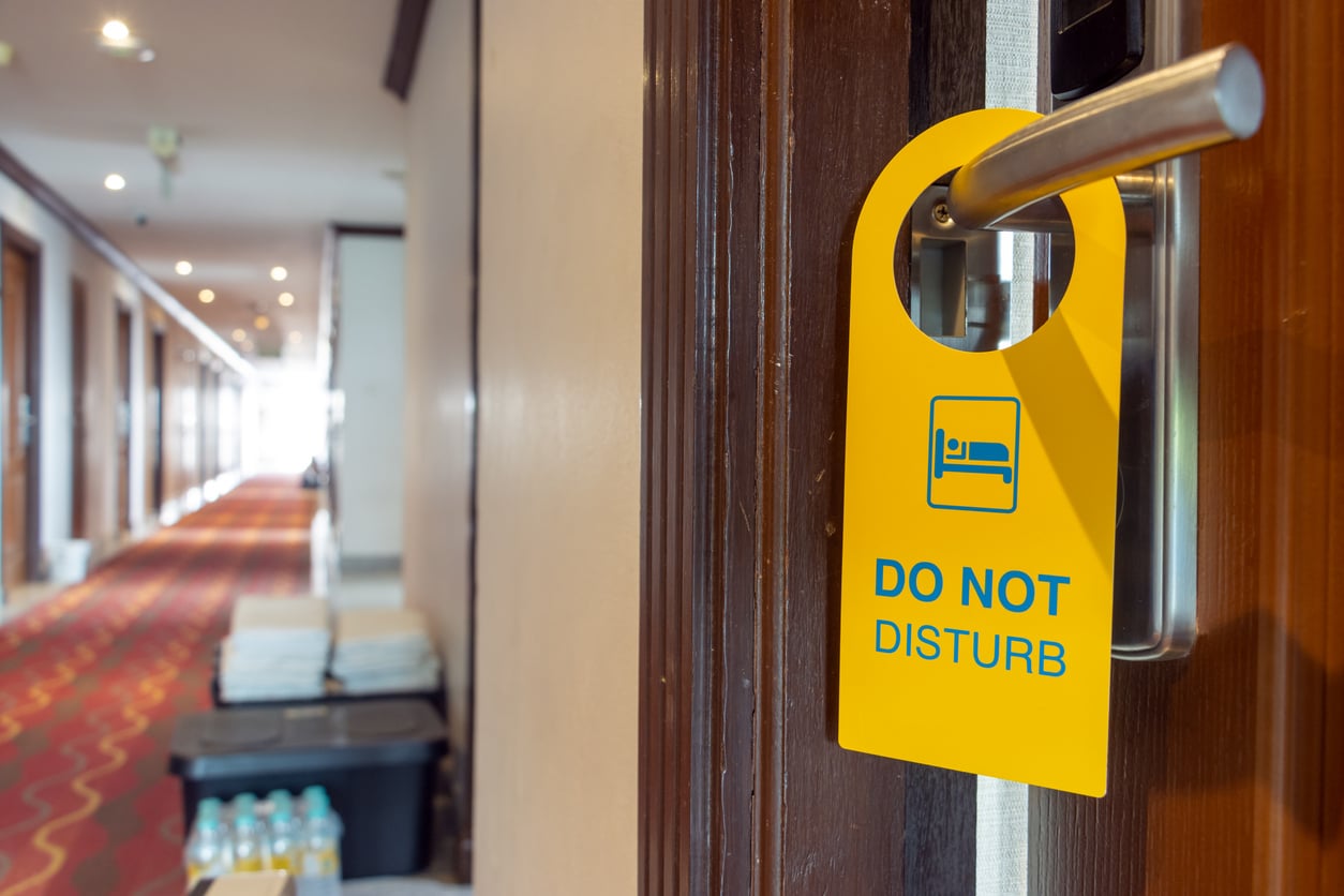 A yellow "Do Not Disturb" sign hangs on a hotel room door handle, emphasizing hotel room safety, with a long hallway and cleaning supplies visible in the background.