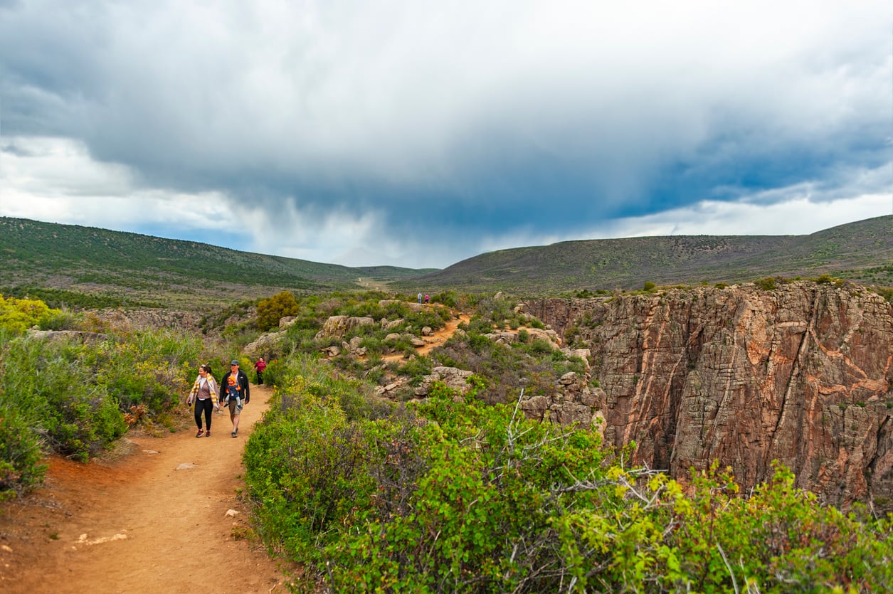 Hikers walk along a dirt path through green shrubland beside a rocky canyon under a cloudy sky, with distant hills in the background.