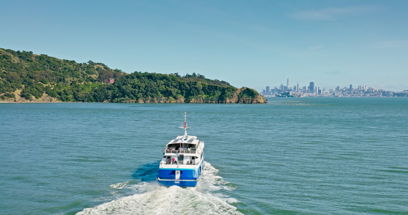 A blue and white ferry travels across a bay toward a green, tree-covered island, with a distant city skyline visible under a clear blue sky.