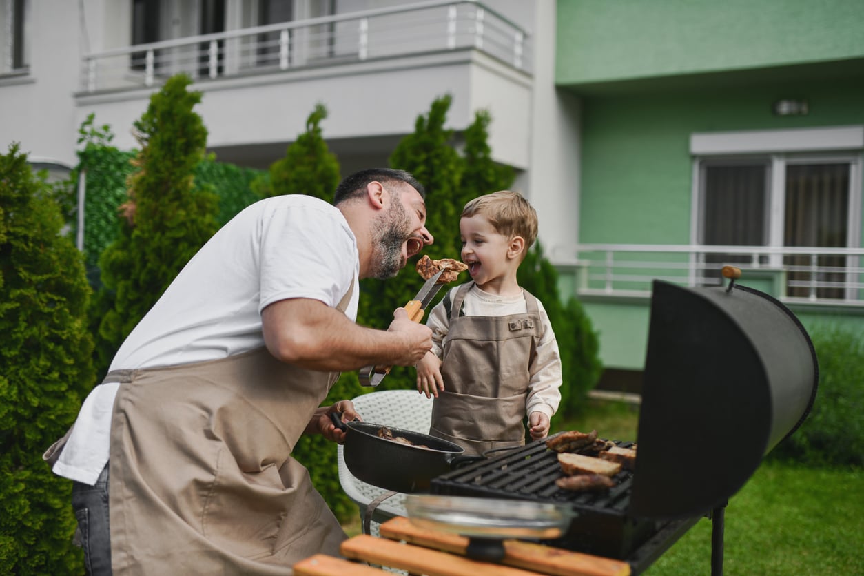 A man and a young child, both wearing aprons, smile and laugh as they share a piece of grilled meat on a skewer—enjoying timeless barbecue moments with classic products men still buy—while cooking together in the backyard.