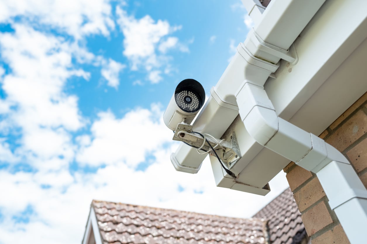 A security camera mounted on the corner of a building's roof, with white gutters and brick walls visible. The background shows a blue sky with scattered clouds and part of a tiled roof.