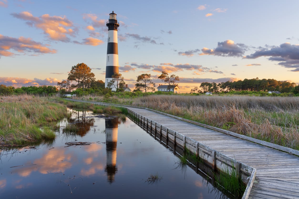 A black and white striped lighthouse stands near a marsh, reflected in calm water alongside a wooden boardwalk at sunset, offering one of the many scenic Florida alternatives with trees and scattered clouds in the sky.