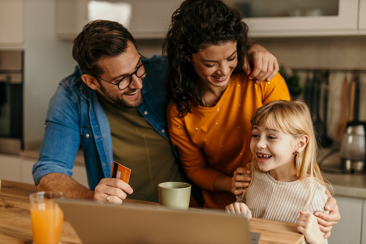 A man, woman, and young girl smile and look at a laptop together in a kitchen. The man holds a credit card, suggesting they are shopping or making a payment online. A mug and a glass of orange juice sit on the table.