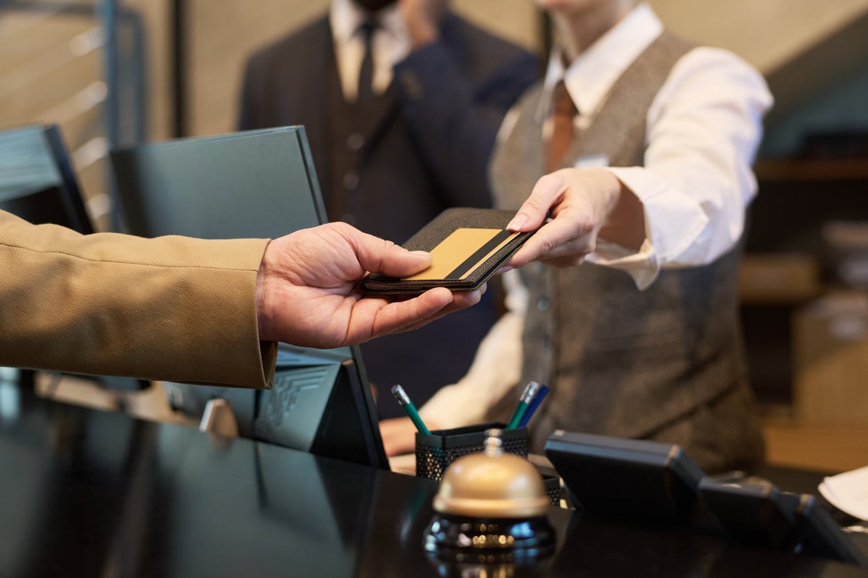 A hotel receptionist in uniform hands a passport and a gold card back to a guest across the front desk, with other staff and office supplies visible in the background.