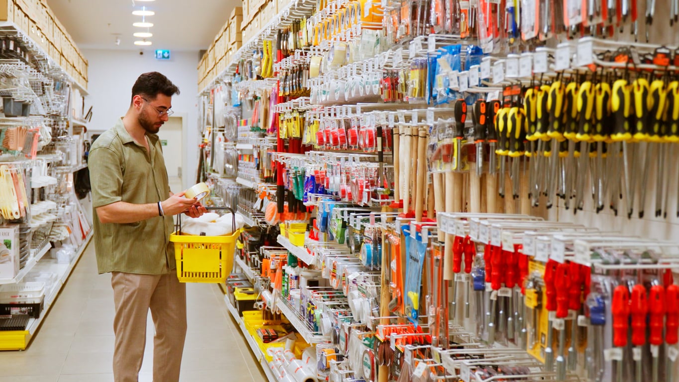 A man holding a yellow shopping basket examines products men still buy in an aisle filled with various tools and hardware supplies at a store.