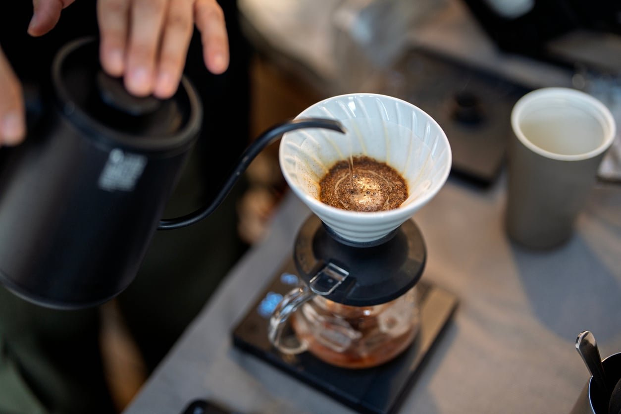 A person pours hot water from a black kettle over ground coffee in a white dripper, making pour-over coffee. The setup includes a glass carafe, a digital scale, and a paper coffee cup on a table.